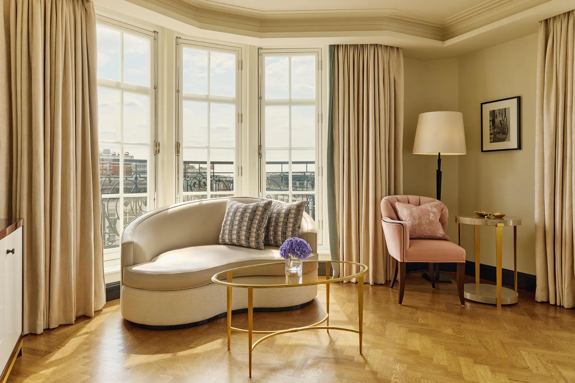 Elegant sitting area with a curved cream sofa, pink armchair, and gold coffee tables beside tall bay windows framed by beige curtains, overlooking city rooftops.