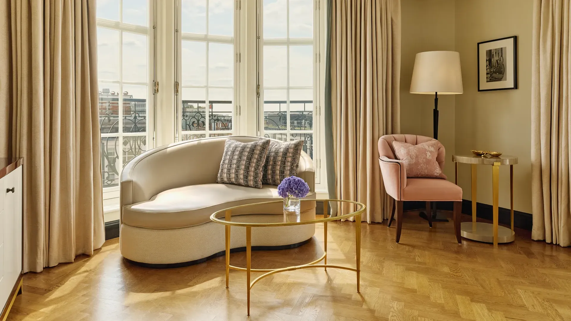 Elegant sitting area with a curved cream sofa, pink armchair, and gold coffee tables beside tall bay windows framed by beige curtains, overlooking city rooftops.