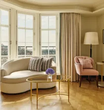Elegant sitting area with a curved cream sofa, pink armchair, and gold coffee tables beside tall bay windows framed by beige curtains, overlooking city rooftops.