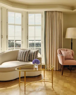 Elegant sitting area with a curved cream sofa, pink armchair, and gold coffee tables beside tall bay windows framed by beige curtains, overlooking city rooftops.