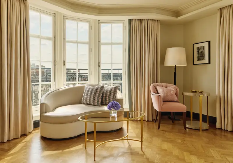 Elegant sitting area with a curved cream sofa, pink armchair, and gold coffee tables beside tall bay windows framed by beige curtains, overlooking city rooftops.