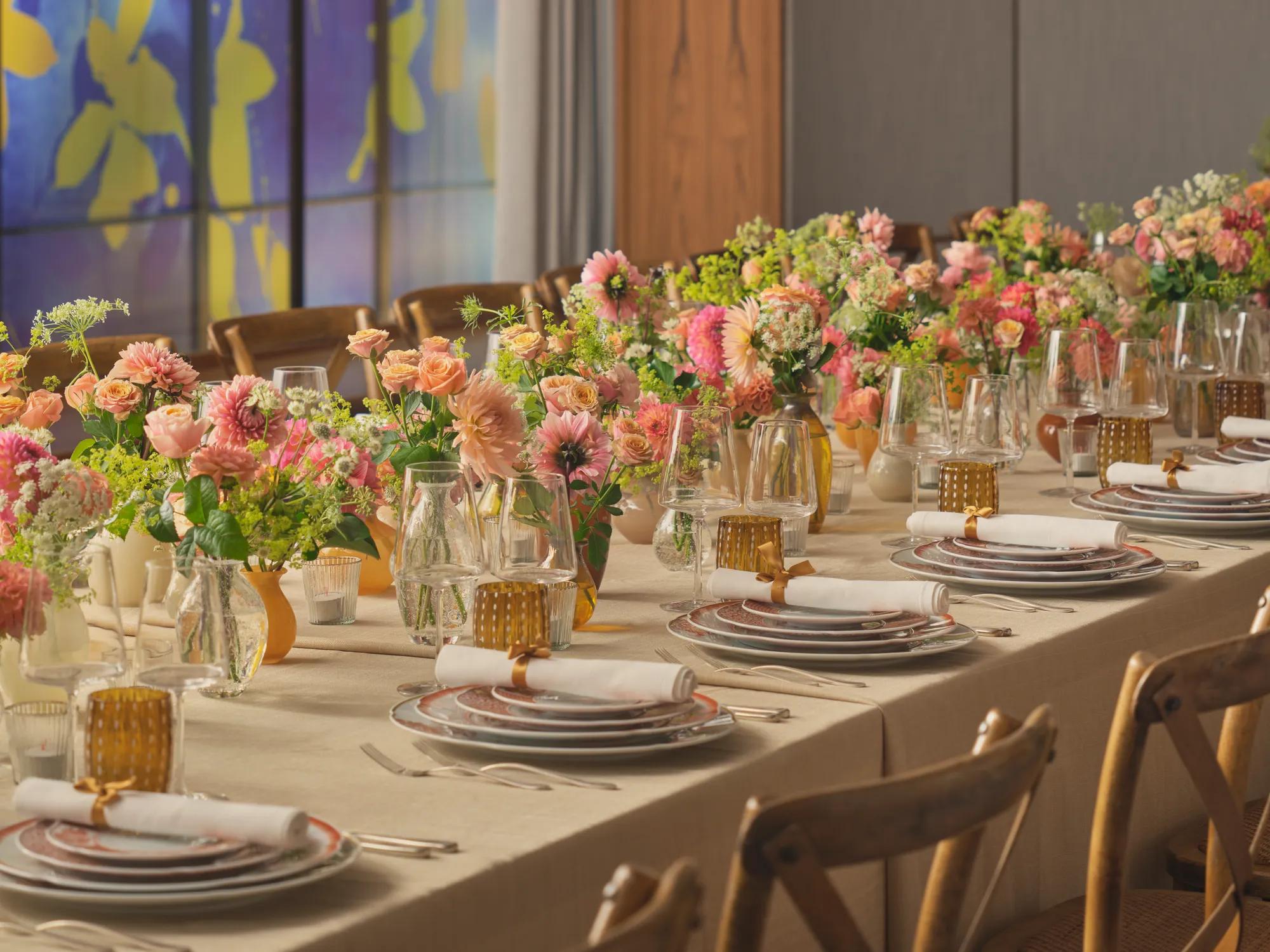Long dining table set for an event, decorated with pink and peach flowers and glowing amber glassware