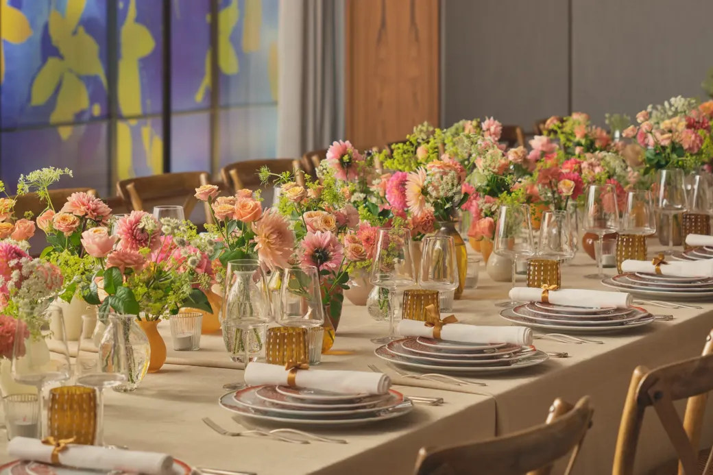 Long dining table set for an event, decorated with pink and peach flowers and glowing amber glassware