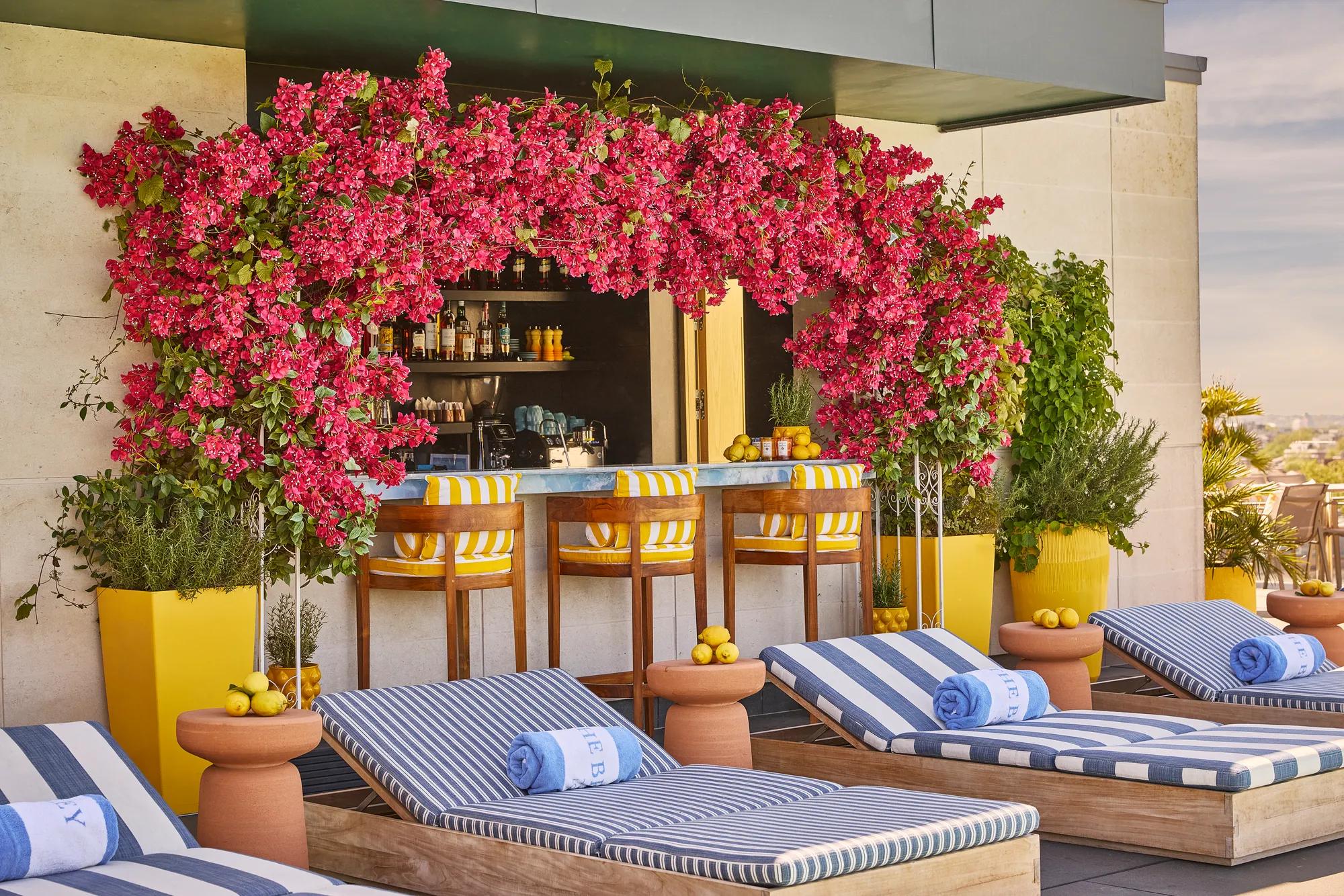 Rooftop poolside bar framed by bright pink flowers, with striped loungers, rolled towels and yellow accents.