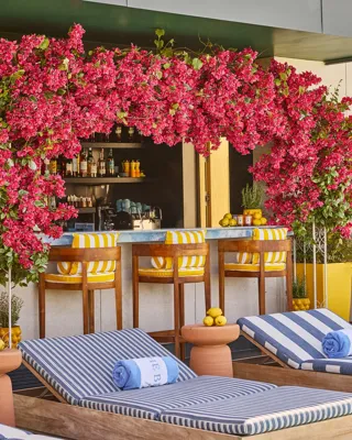 Rooftop poolside bar framed by bright pink flowers, with striped loungers, rolled towels and yellow accents.