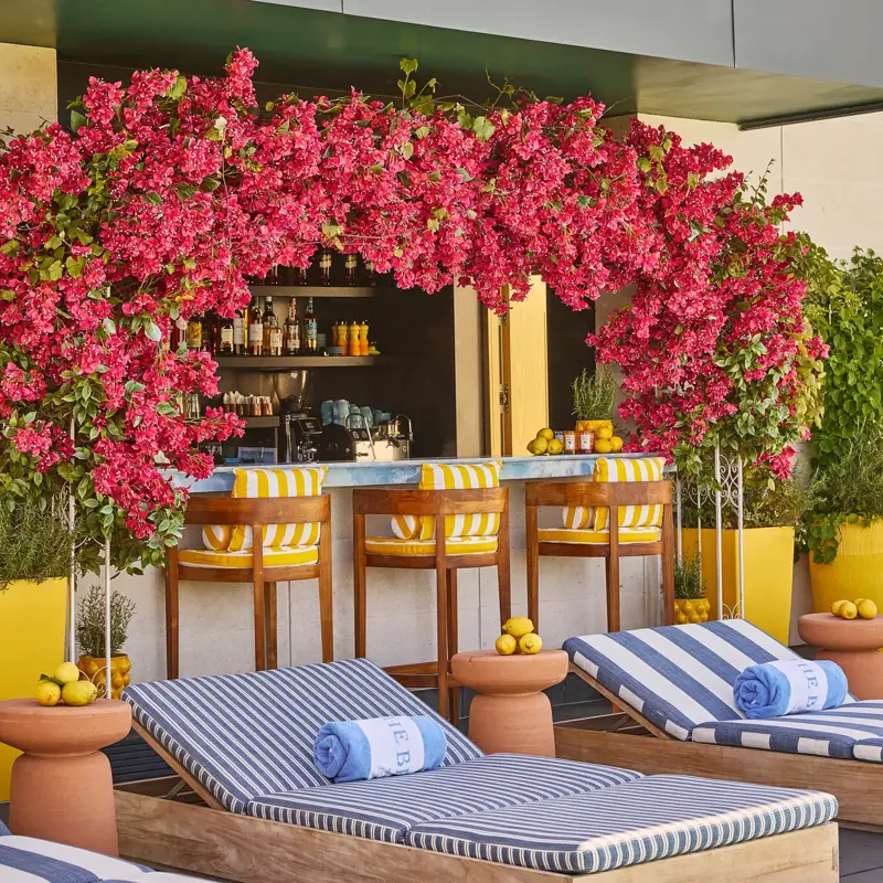 Rooftop poolside bar framed by bright pink flowers, with striped loungers, rolled towels and yellow accents.