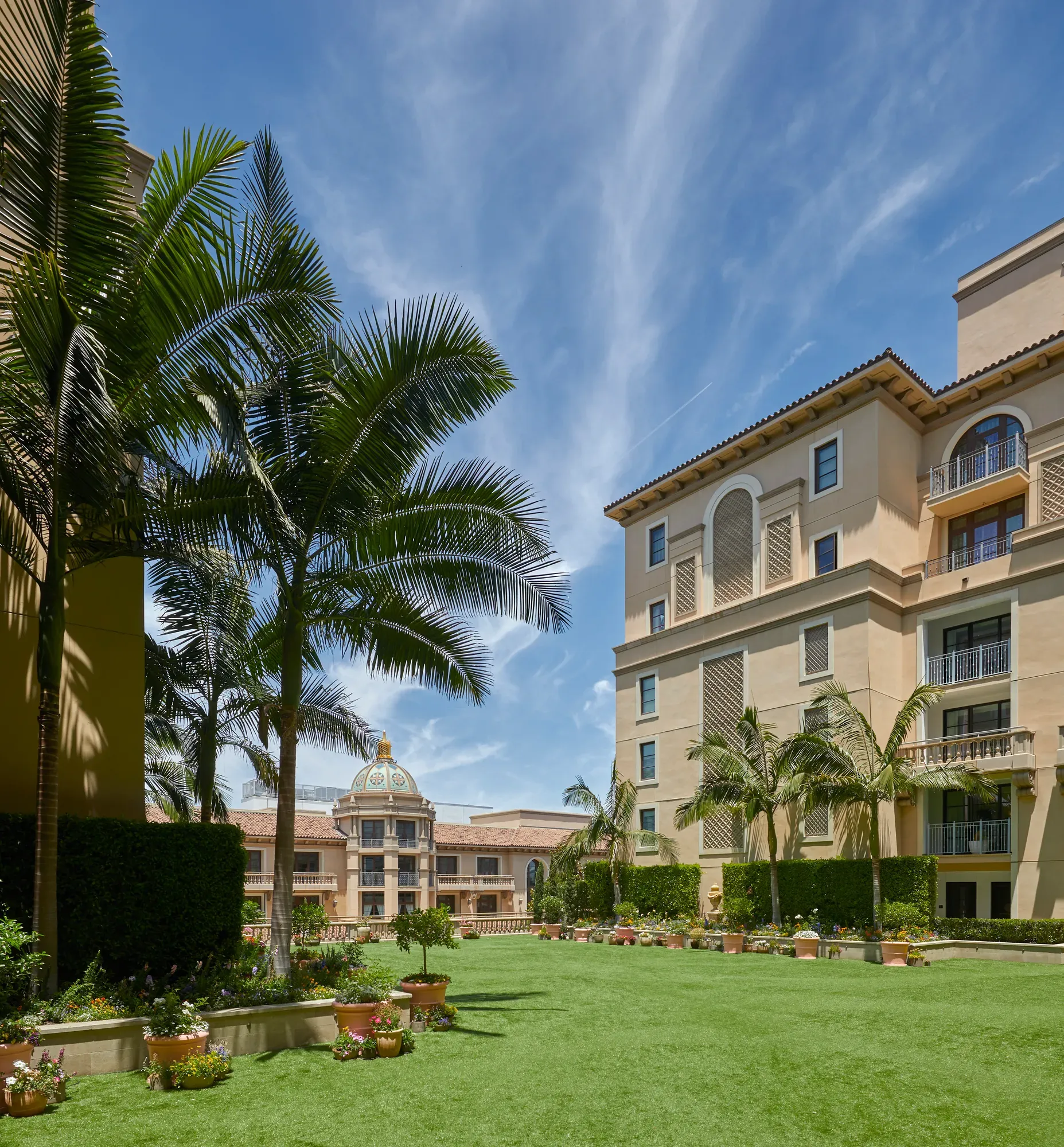 Wide lawn of the Garden Terrace bordered by palms, potted flowers and hotel façades beneath a bright, streaked blue sky.