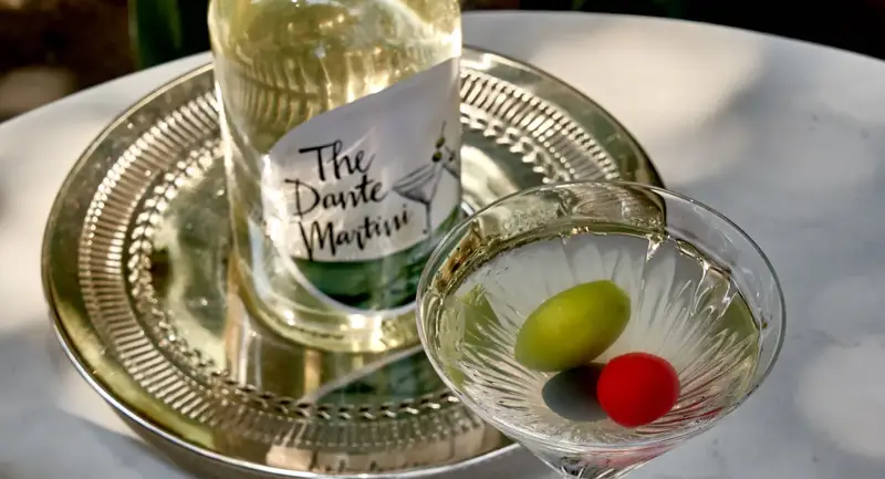 Martini glass with green and red olives beside bottled Dante Martini.