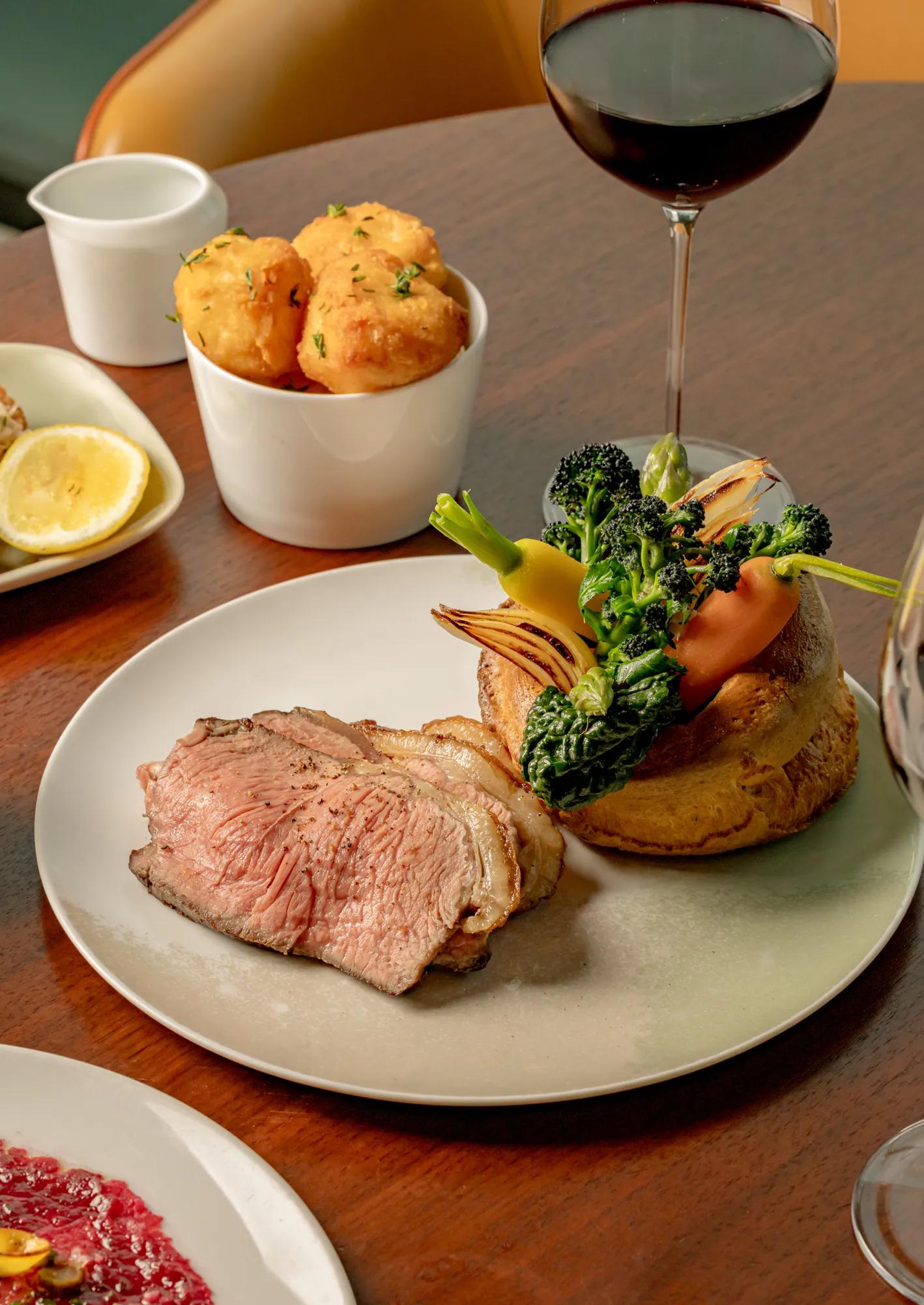 Roast dry-aged beef served with a golden Yorkshire pudding, seasonal green vegetables and roasted shallots on a ceramic plate, accompanied by red wine and side dishes on a wooden dining table.