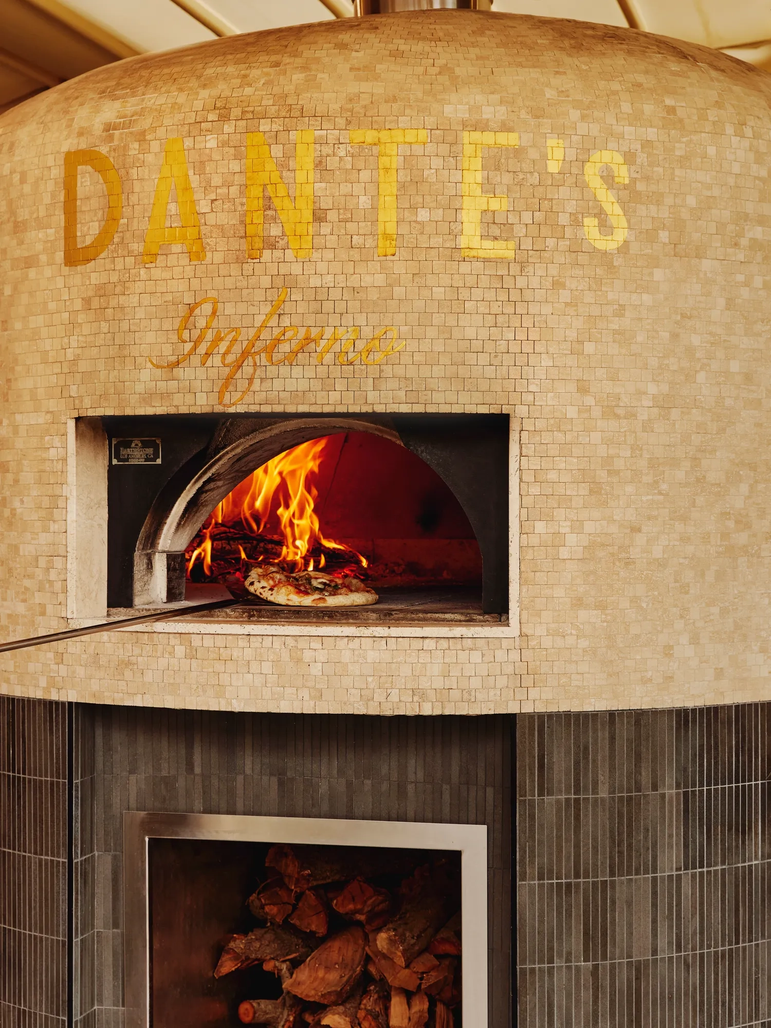 Gold-tiled wood-fired oven labelled “DANTE’S Inferno” with a pizza baking inside and stacked firewood below.