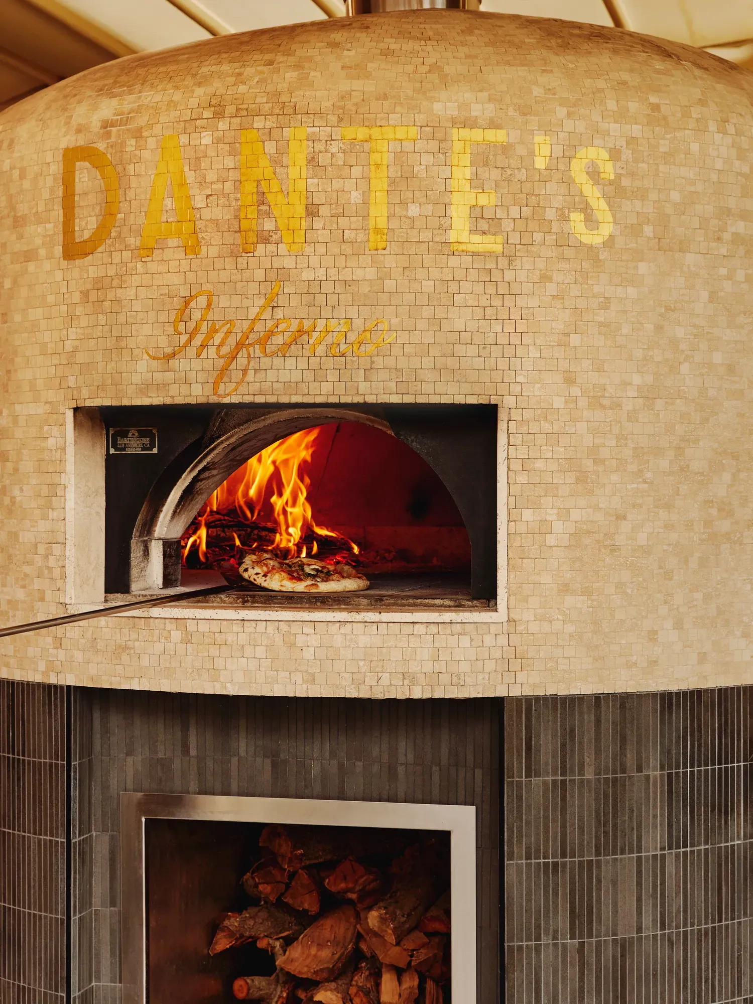 Gold-tiled wood-fired oven labelled “DANTE’S Inferno” with a pizza baking inside and stacked firewood below.