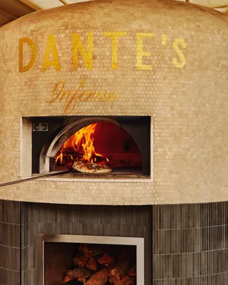 Gold-tiled wood-fired oven labelled “DANTE’S Inferno” with a pizza baking inside and stacked firewood below.