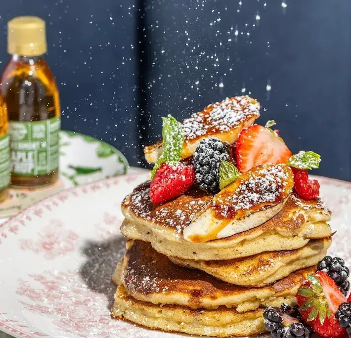 Stack of fluffy pancakes topped with berries and bananas, dusted with powdered sugar.