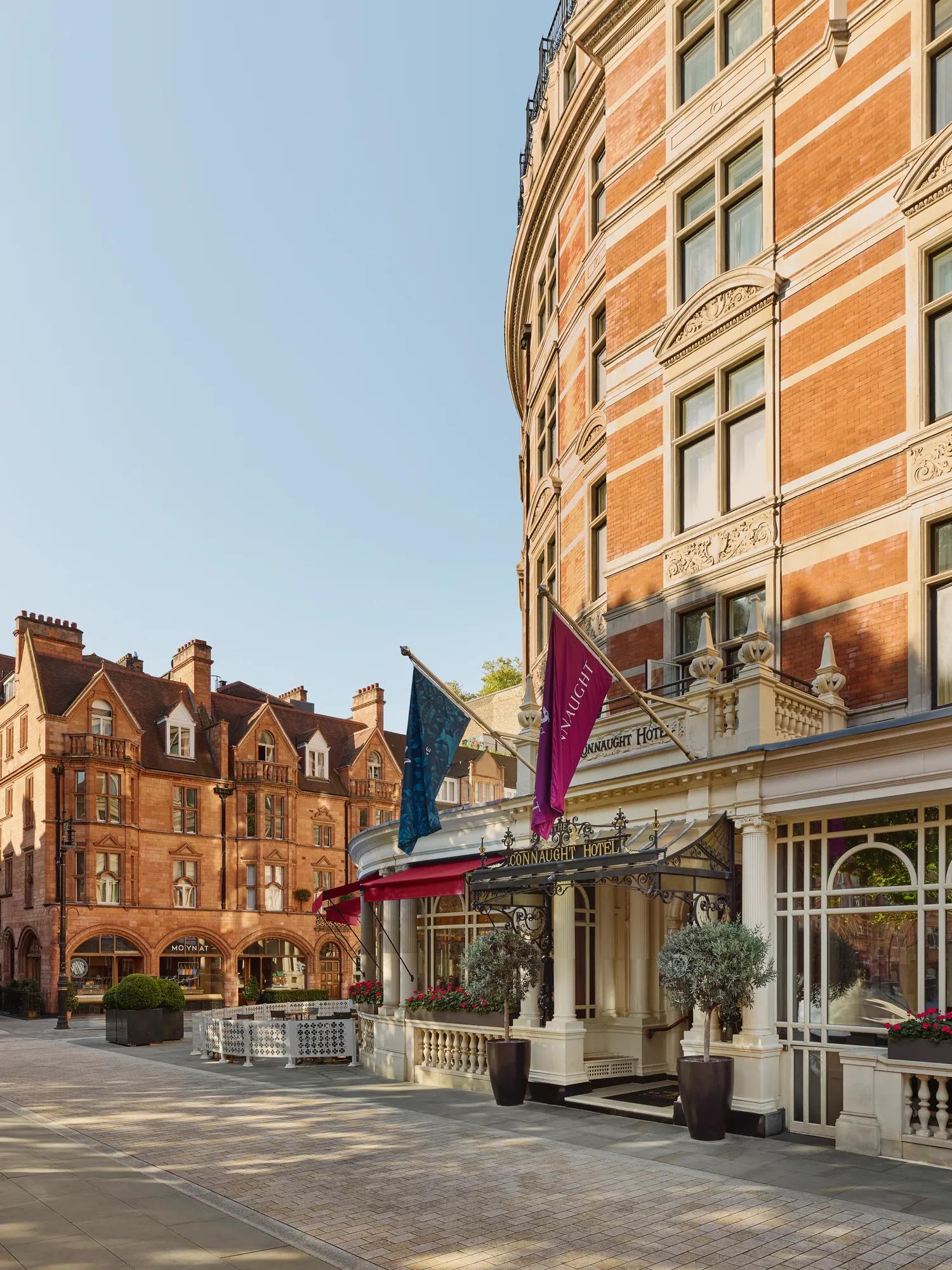 Exterior of The Connaught hotel in Mayfair, London, showing a curved red-brick façade with cream stone detailing, three flags over the entrance canopy, and potted trees by the doorway, with adjacent period buildings under a clear sky.