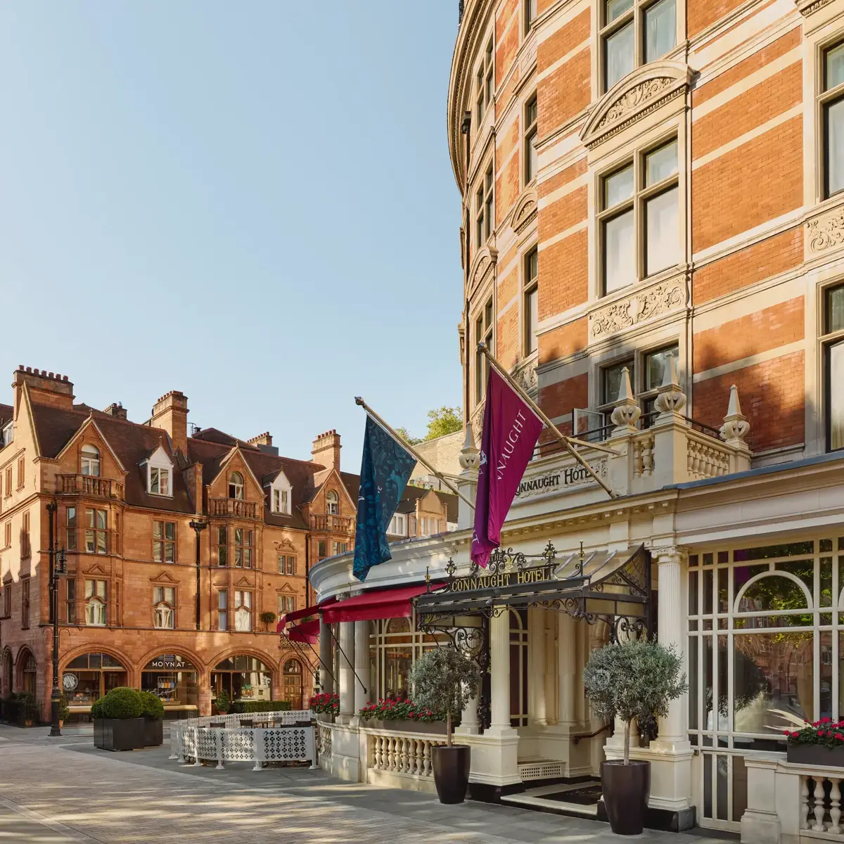Exterior of The Connaught hotel in Mayfair, London, showing a curved red-brick façade with cream stone detailing, three flags over the entrance canopy, and potted trees by the doorway, with adjacent period buildings under a clear sky.
