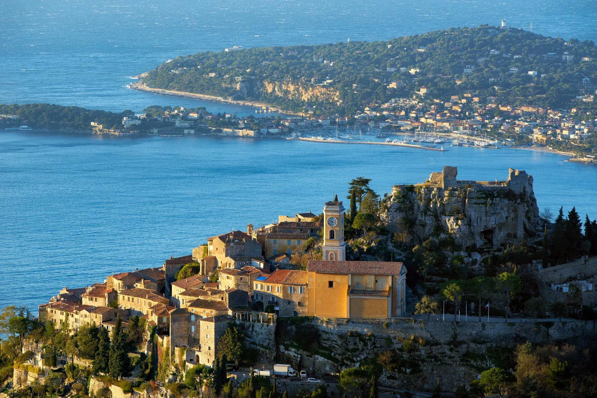 Village perché avec des maisons en pierre et un clocher dominant la mer, accompagné d’une ruine rocheuse voisine et d’une ville côtière visible de l’autre côté de la baie.