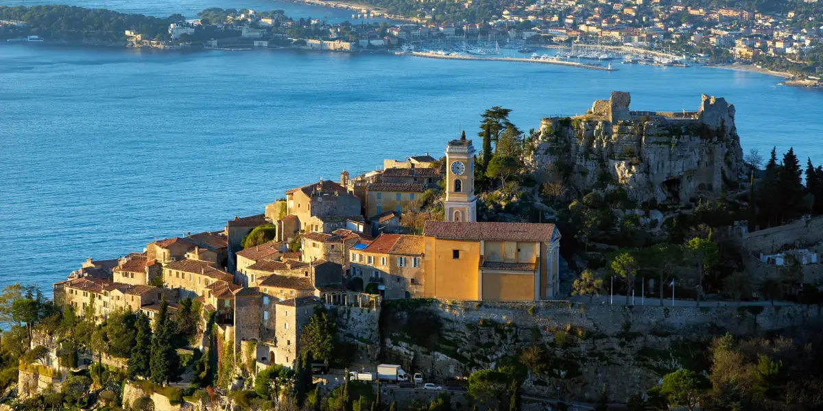 Hilltop village with stone houses and a church tower overlooking the sea, with a rocky ruin nearby and a coastal town visible across the bay.