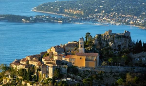 Village perché avec des maisons en pierre et un clocher dominant la mer, accompagné d’une ruine rocheuse voisine et d’une ville côtière visible de l’autre côté de la baie.