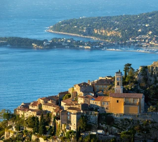 Hilltop village with stone houses and a church tower overlooking the sea, with a rocky ruin nearby and a coastal town visible across the bay.