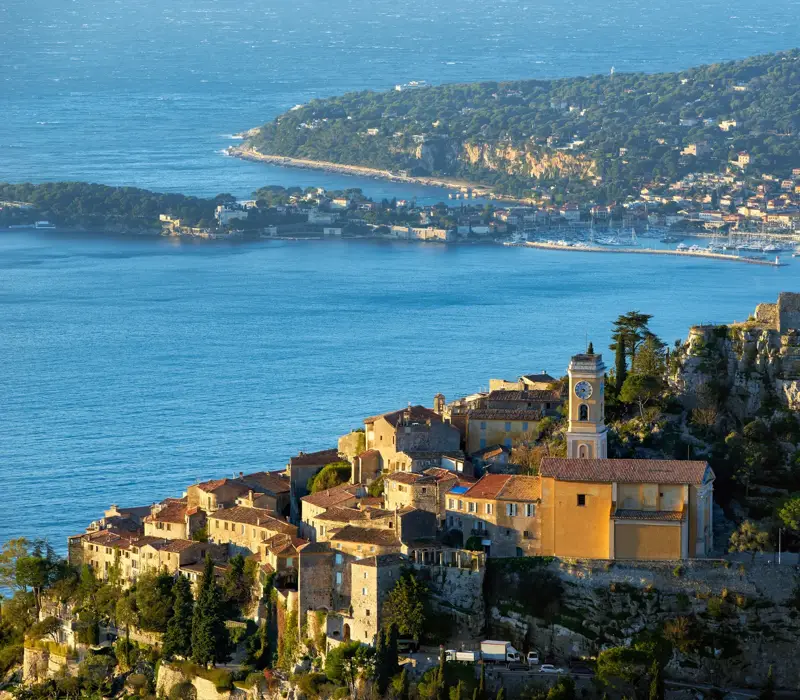 Hilltop village with stone houses and a church tower overlooking the sea, with a rocky ruin nearby and a coastal town visible across the bay.