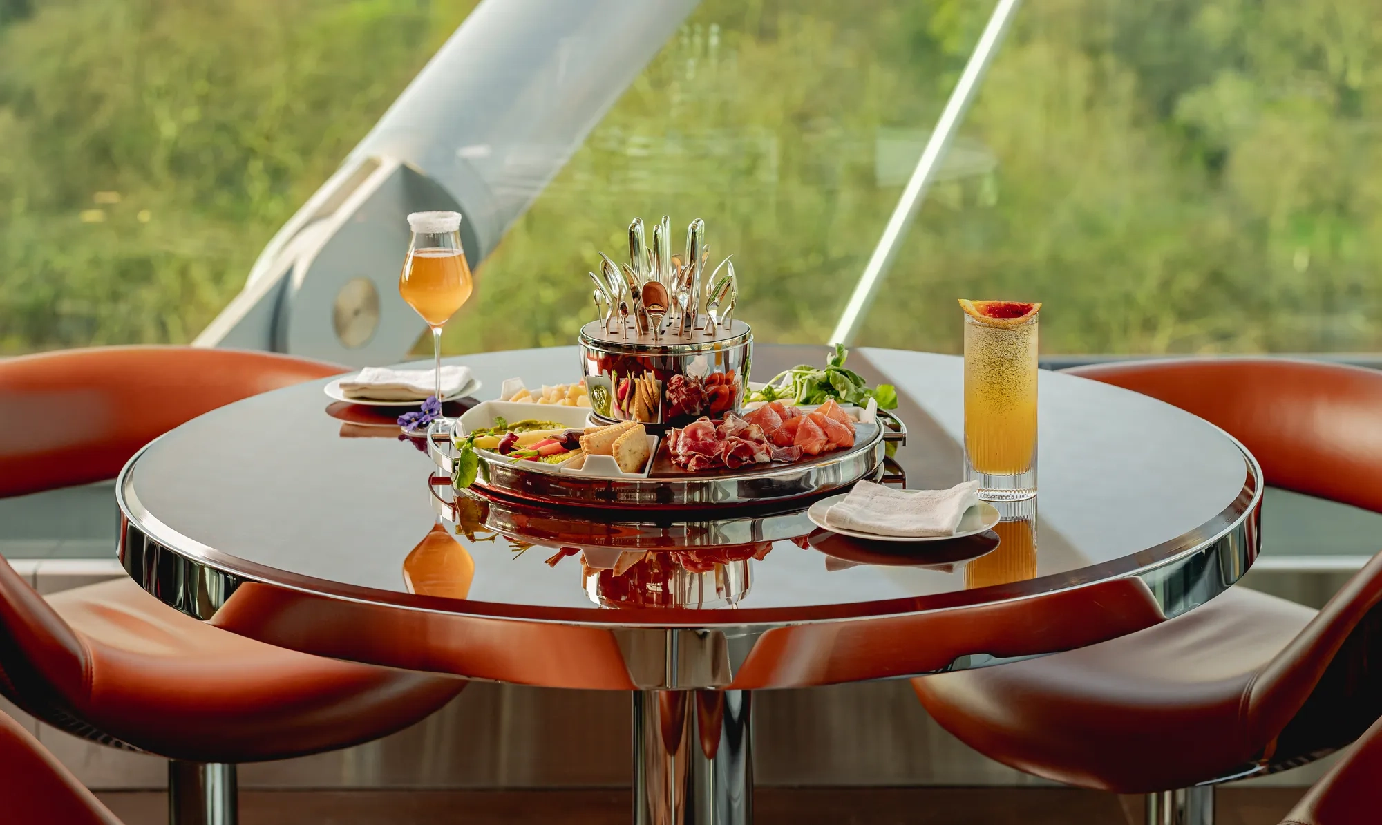 Round table set with charcuterie platter and cocktails, surrounded by leather chairs and overlooking greenery through large windows.