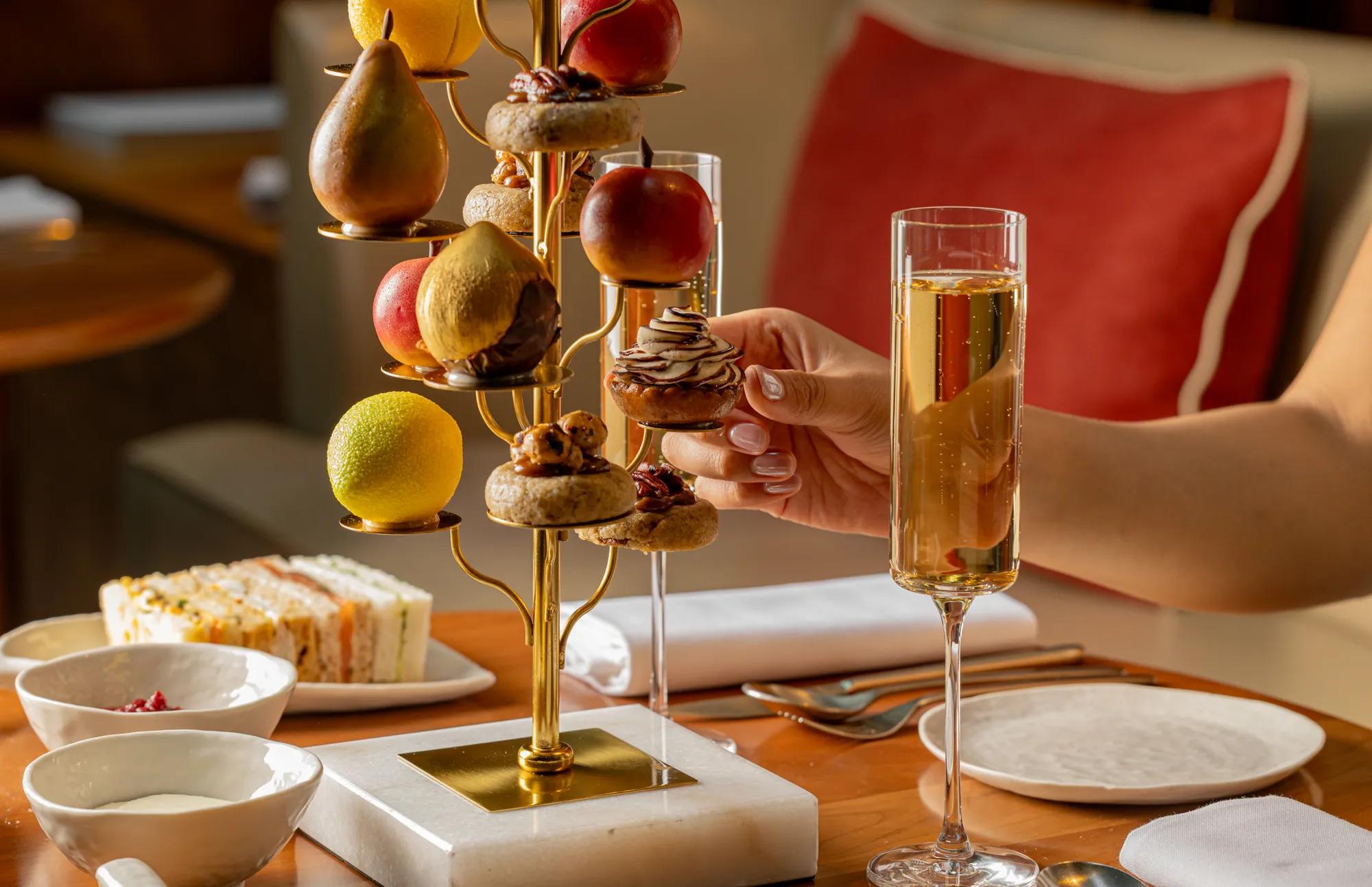 Hand selecting a pastry from a fruit-inspired afternoon tea stand, served with sandwiches and sparkling wine.