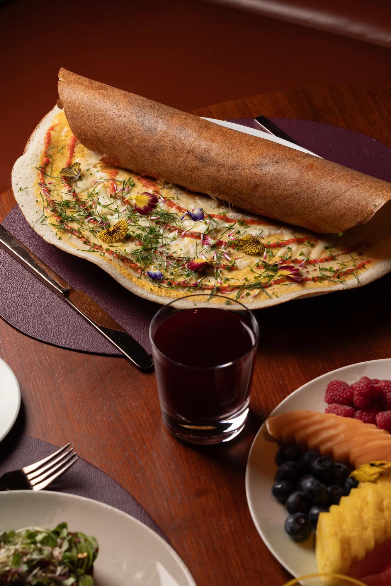 Large dosa topped with edible flowers and herbs, served with juice, salad, and fresh fruit.