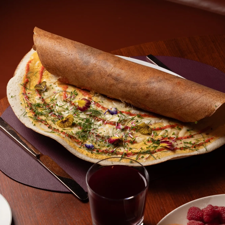 Large dosa topped with edible flowers and herbs, served with juice, salad, and fresh fruit.