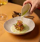 Hand pouring green sauce over plated gourmet dish, with wine glass and cutlery on wooden table.