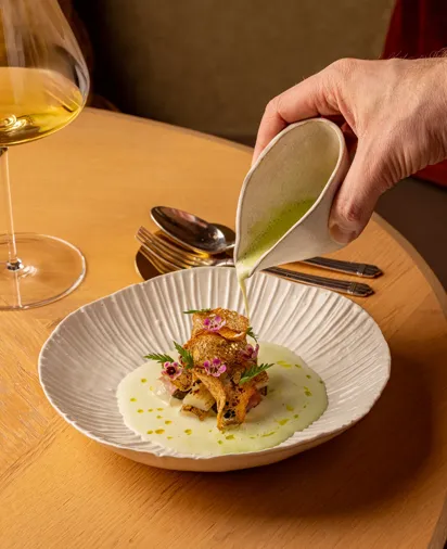 Hand pouring green sauce over plated gourmet dish, with wine glass and cutlery on wooden table.