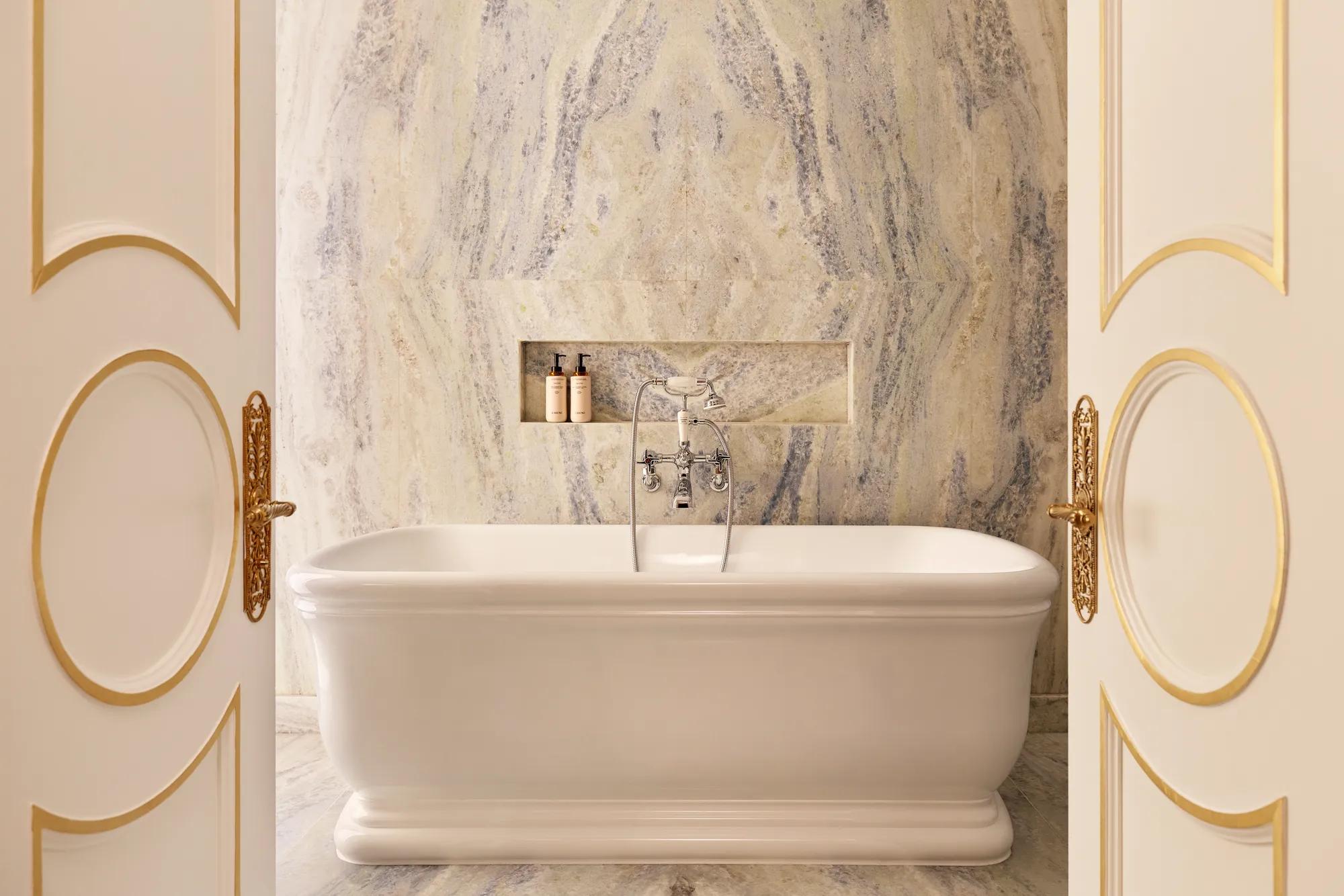 Freestanding bathtub set before a marble wall with built-in niche and chrome fixtures, framed by open double doors with gold detailing.