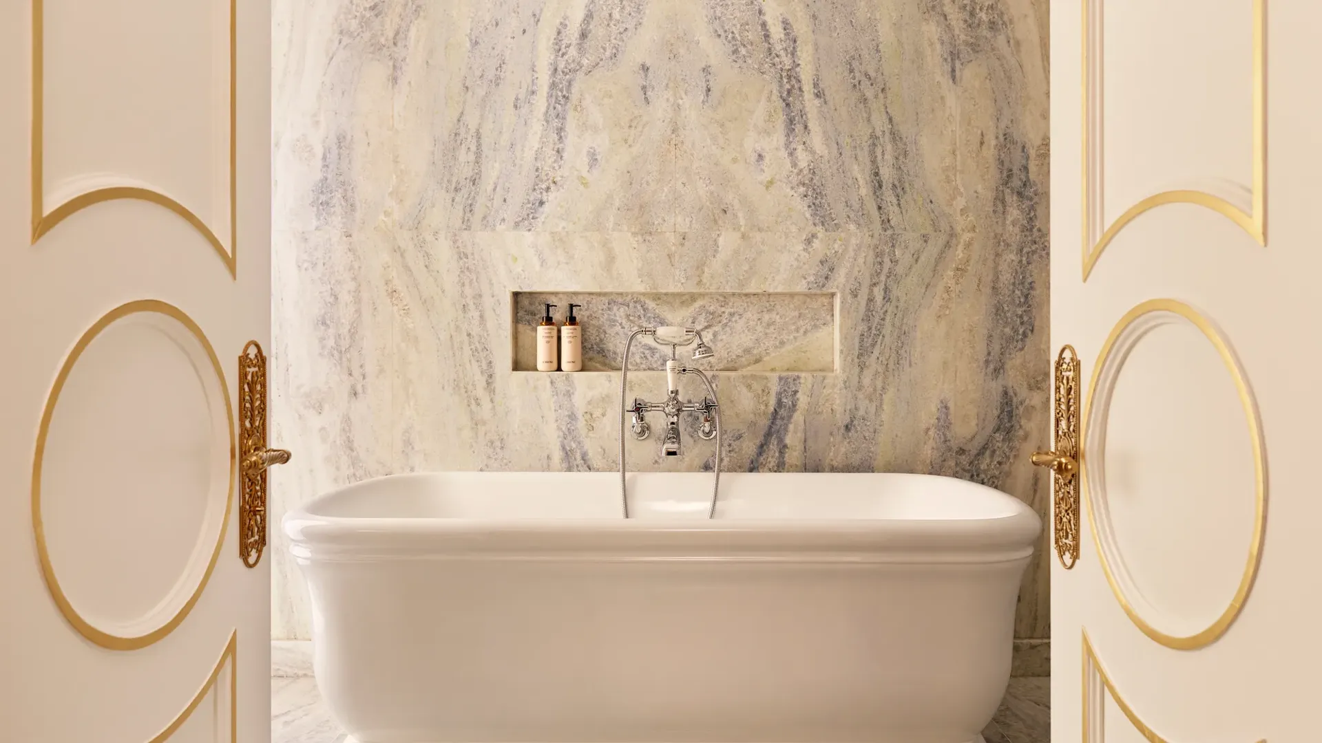Freestanding bathtub set before a marble wall with built-in niche and chrome fixtures, framed by open double doors with gold detailing.