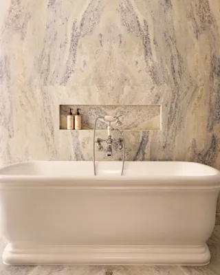 Freestanding bathtub set before a marble wall with built-in niche and chrome fixtures, framed by open double doors with gold detailing.