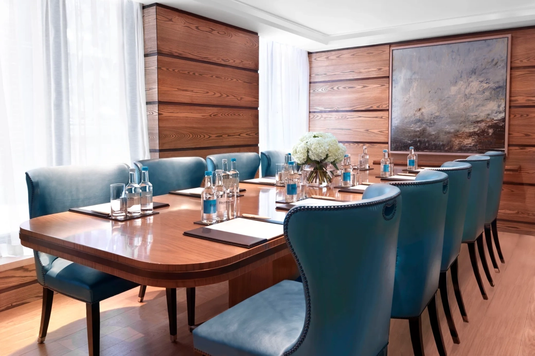 Bright meeting room with a polished wooden boardroom table, blue leather chairs, and bottled water set for attendees.