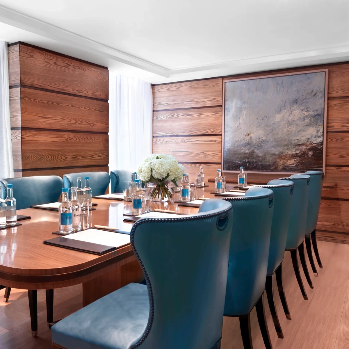Bright meeting room with a polished wooden boardroom table, blue leather chairs, and bottled water set for attendees.