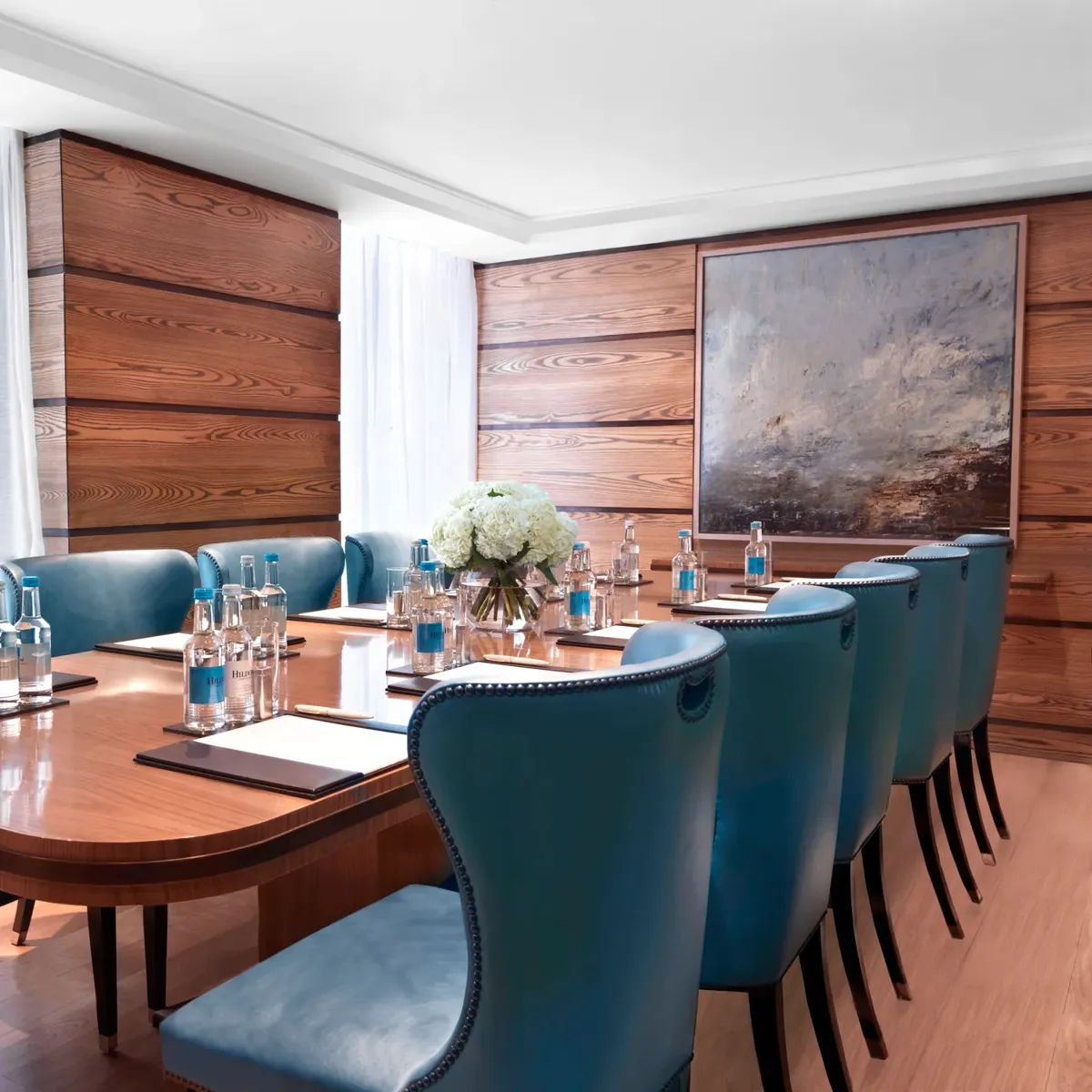 Bright meeting room with a polished wooden boardroom table, blue leather chairs, and bottled water set for attendees.