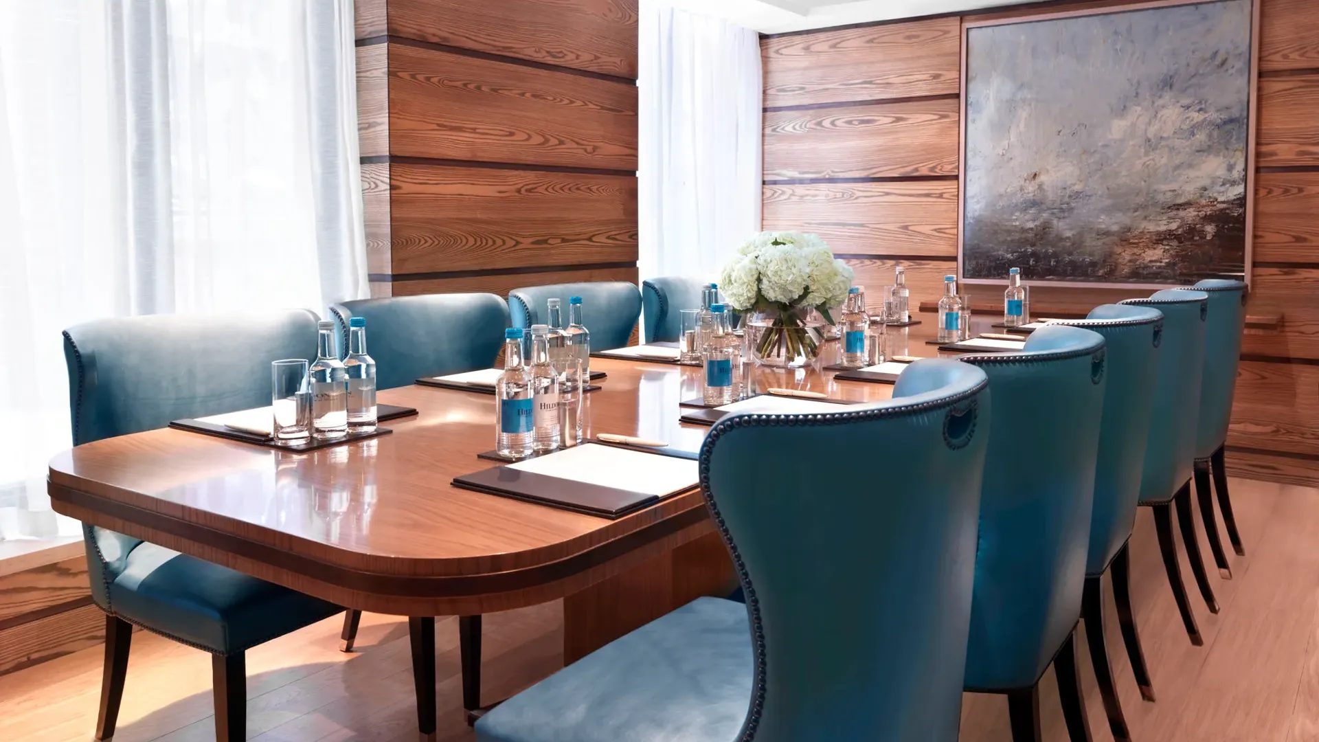 Bright meeting room with a polished wooden boardroom table, blue leather chairs, and bottled water set for attendees.