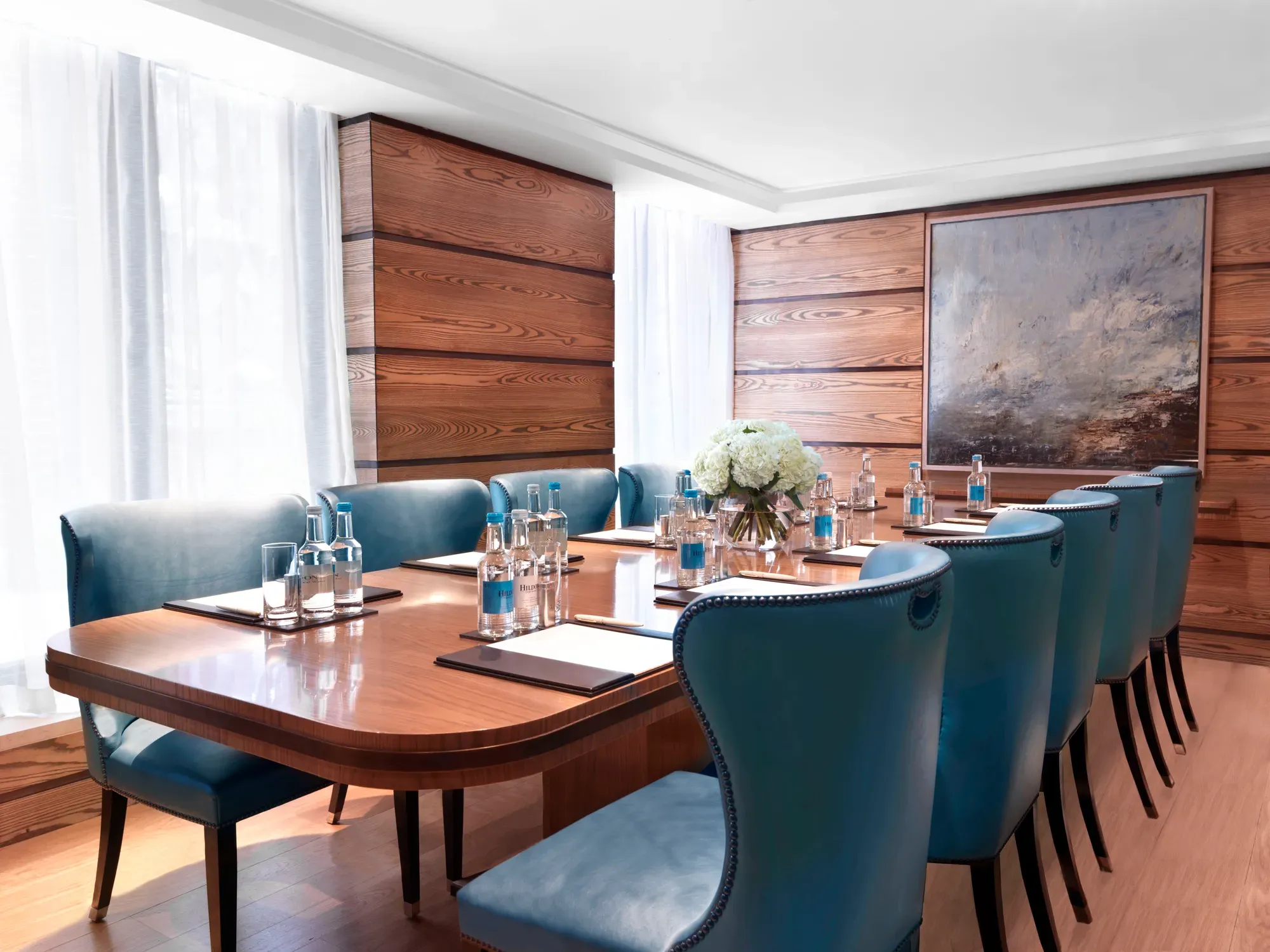 Bright meeting room with a polished wooden boardroom table, blue leather chairs, and bottled water set for attendees.