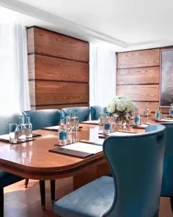 Bright meeting room with a polished wooden boardroom table, blue leather chairs, and bottled water set for attendees.