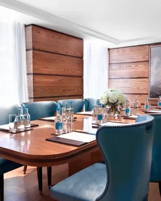 Bright meeting room with a polished wooden boardroom table, blue leather chairs, and bottled water set for attendees.