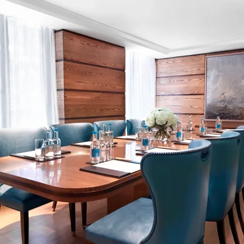 Bright meeting room with a polished wooden boardroom table, blue leather chairs, and bottled water set for attendees.