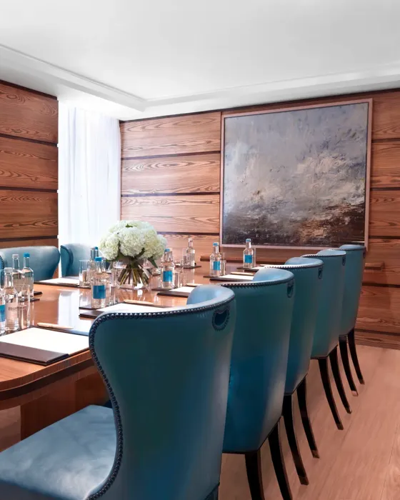 Bright meeting room with a polished wooden boardroom table, blue leather chairs, and bottled water set for attendees.