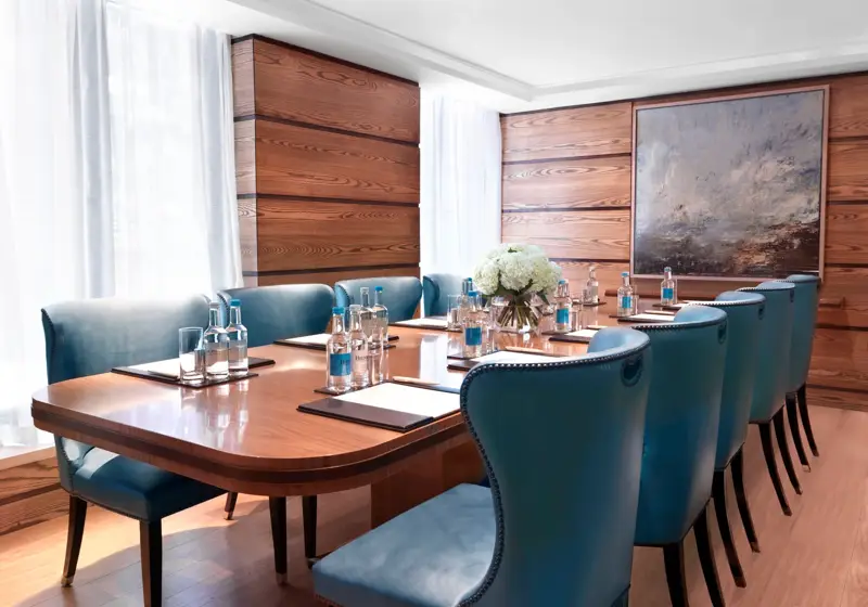 Bright meeting room with a polished wooden boardroom table, blue leather chairs, and bottled water set for attendees.