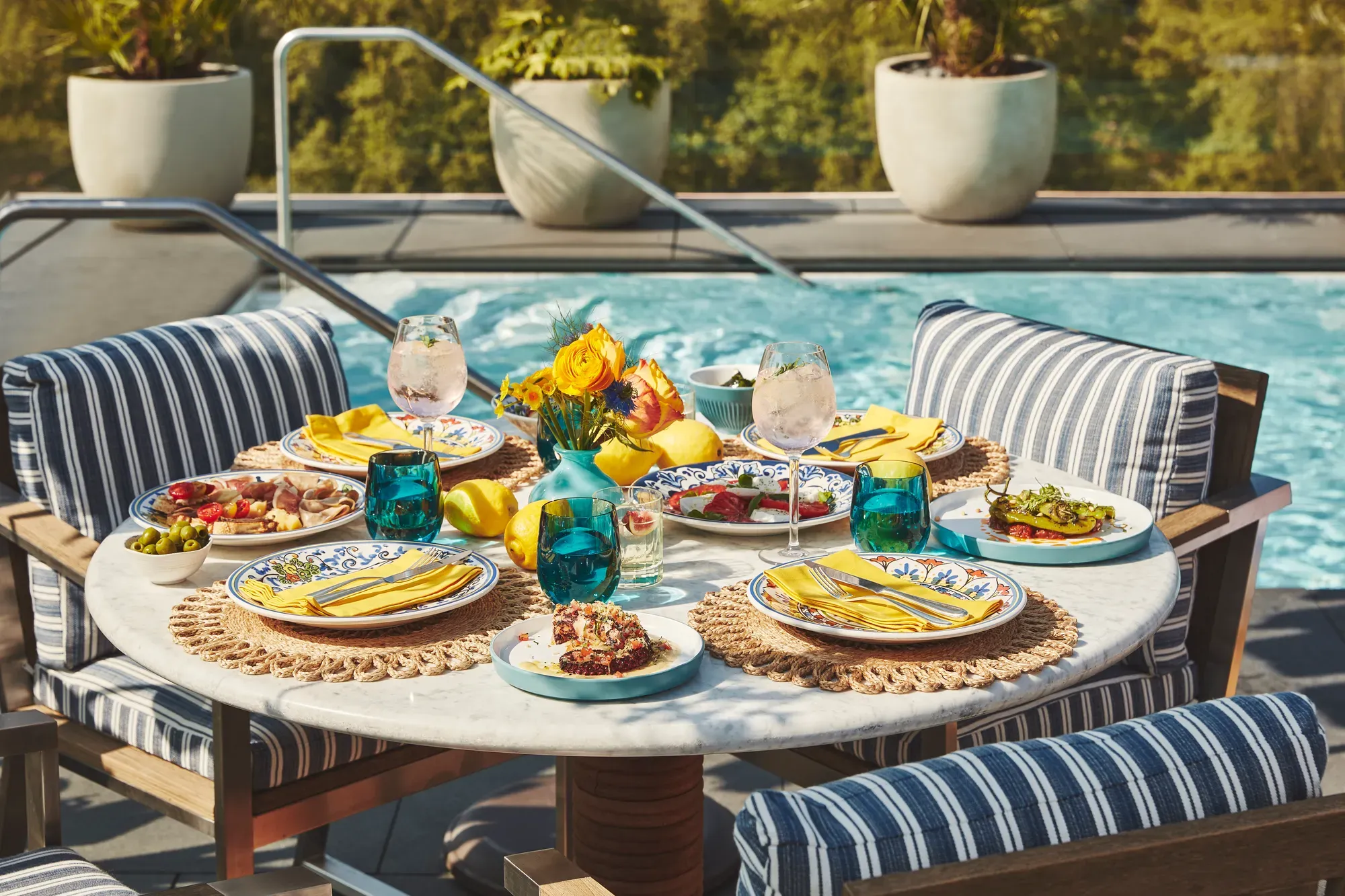 Poolside table set with vibrant dishes, drinks, and yellow napkins surrounded by striped cushioned chairs and greenery.