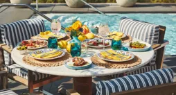 Poolside table set with vibrant dishes, drinks, and yellow napkins surrounded by striped cushioned chairs and greenery.