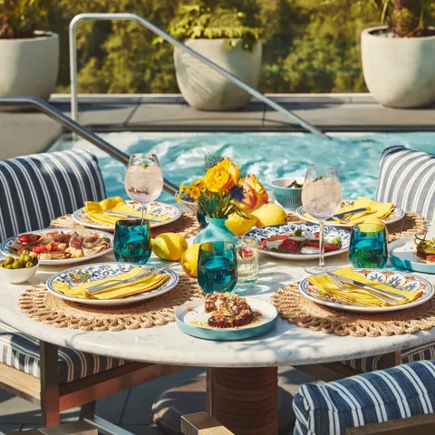 Poolside table set with vibrant dishes, drinks, and yellow napkins surrounded by striped cushioned chairs and greenery.