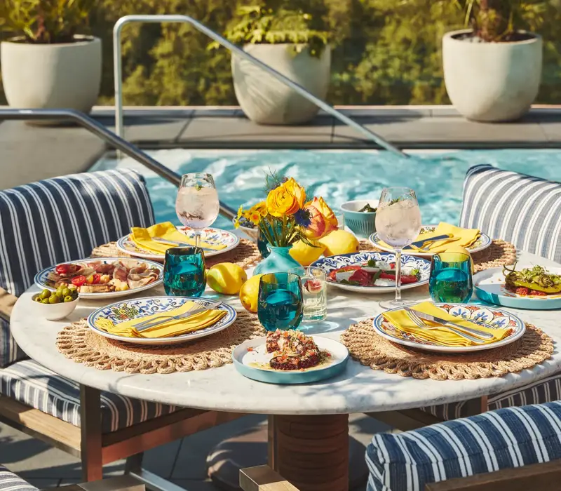 Poolside table set with vibrant dishes, drinks, and yellow napkins surrounded by striped cushioned chairs and greenery.