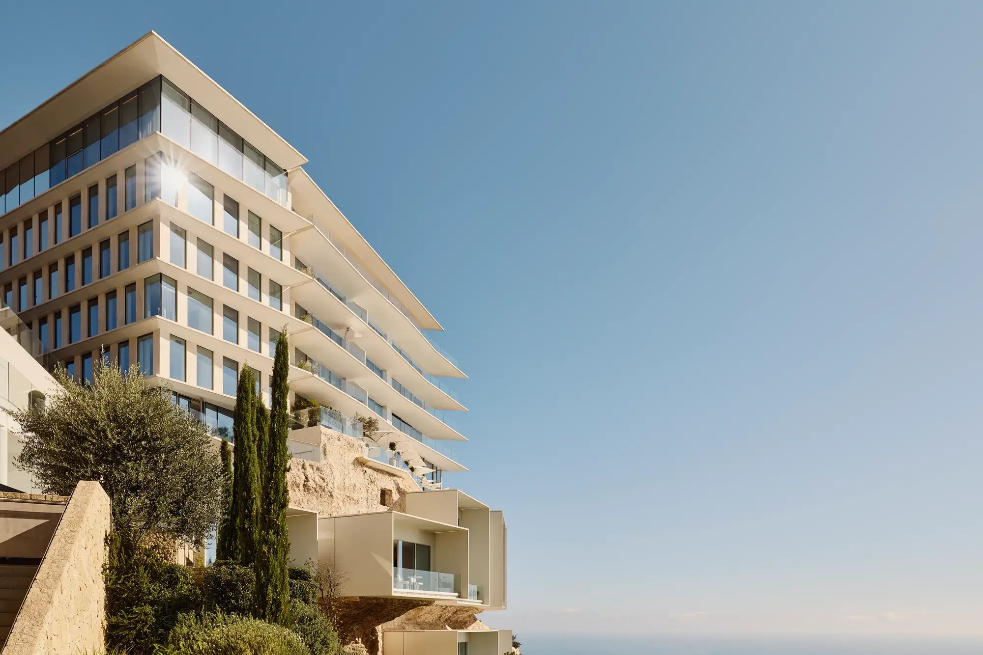 Modern terraced hotel built into a cliffside above the Mediterranean, featuring clean white lines, expansive glass balconies, and panoramic sea views framed by lush coastal greenery.