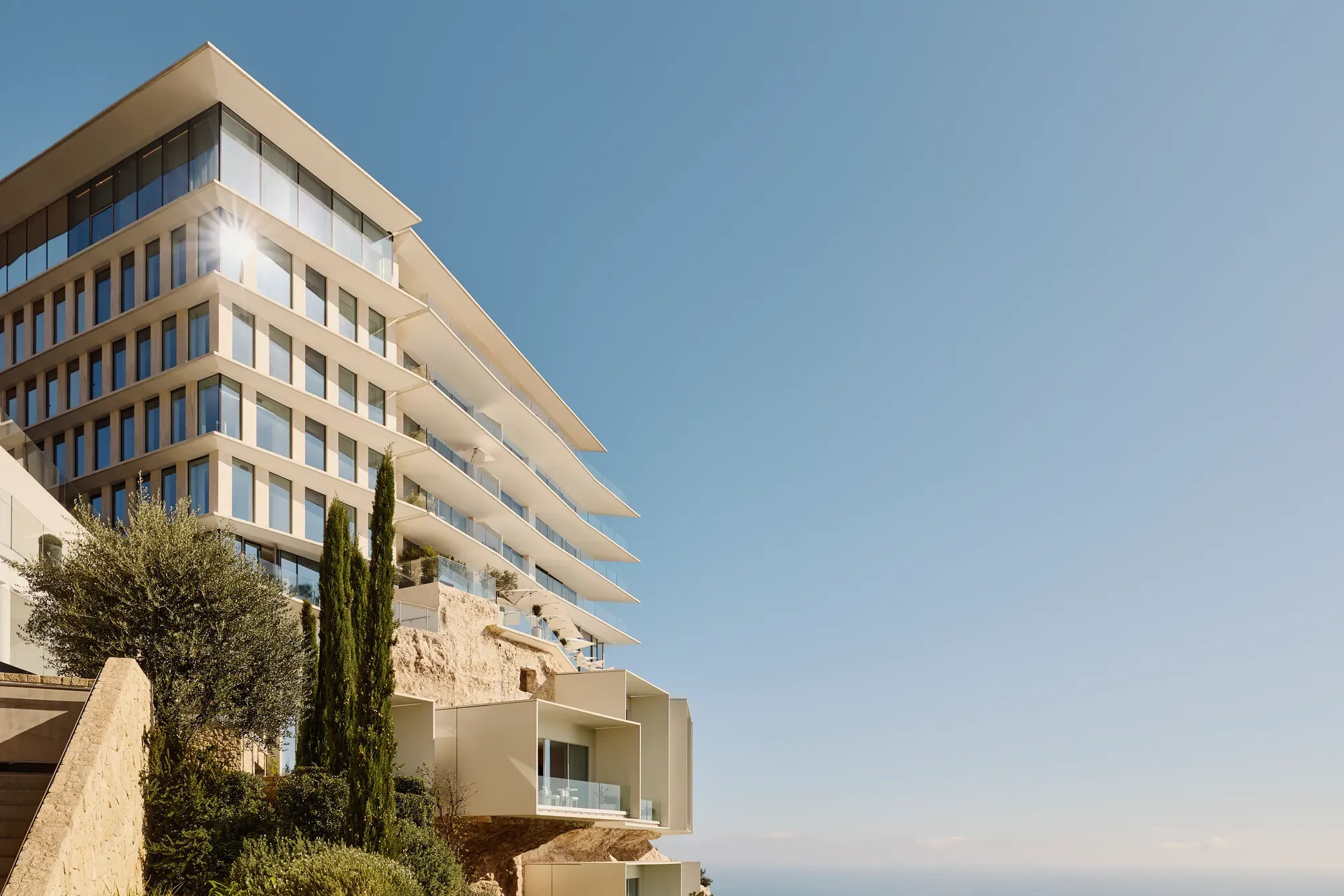 Modern terraced hotel built into a cliffside above the Mediterranean, featuring clean white lines, expansive glass balconies, and panoramic sea views framed by lush coastal greenery.