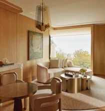 Warm wood-panelled suite living area with curved sofa, round dining table, brass coffee tables, and large window overlooking trees.